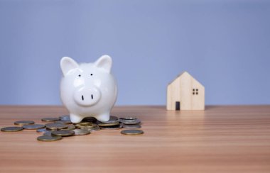 Bir yığın madeni paranın üzerine beyaz bir kumbara yerleştirilir. Ve onun yanında beyaz arka planda ahşap bir model ev var. Hepsi ahşap bir masanın üzerinde.