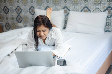 Evde dizüstü bilgisayar kullanan Asyalı bir kadın, rahatlıyor ve uzaktan çalışıyor, modern yaşam tarzı ve teknoloji konsepti, yatak odası ortamında rahat çalışma alanı.