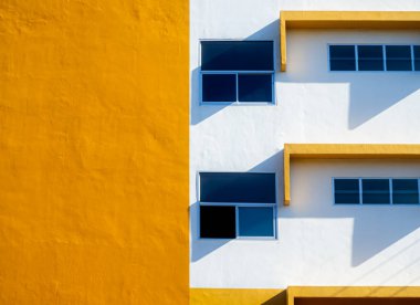 Mavi cam pencereli ve gölgeli modern sarı ve beyaz bina cephesi, minimal mimari arkaplan, güneş ışığı kontrastlı soyut geometrik tasarım.
