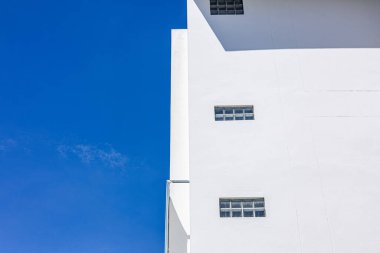 Cam pencereli ve mavi gökyüzü arkaplanlı minimal beyaz bina cephesi, kentsel tasarımda temiz çizgiler ve basitliği gösteren modern mimari konsept.