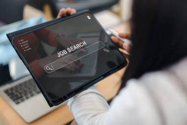 İnternet arayüzü üzerinden iş aramak için dijital tablet kullanan bir kadının, modern ev ofis arka planında dizüstü bilgisayarı olan bir arama çubuğu ve işe alma simgelerinin yer aldığı bir web sitesini kapatın.