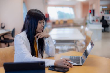 Düşünceli Asyalı iş kadını laptop ekranına bakıyor ve ofiste düşünüyor. Dijital teknoloji ve kablosuz cihazlar kullanarak projede çalışan konsantre kadın profesyonel.