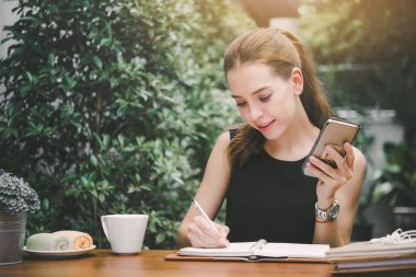 Genç ve güzel bir kadın, arkadaşını cep telefonuyla arar ve bir not alır, bahçede oturur.  