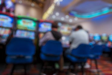 Kumarhanenin içi bulanık iki oyuncunun parlak kumar makinelerinde oturduğunu gösteriyor. Renkli ışıklandırma, parlayan ekranlar ve yumuşak odak etkisi kumar ve gece hayatı temaları için ideal soyut bir eğlence ortamı yaratıyor..