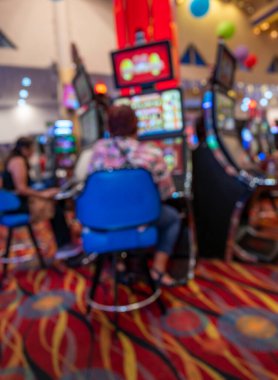 Parlak bir kumarhanenin içinde, renkli ekranlar, desenli halı ve mavi sandalyeler ile canlı, yumuşak odaklı bir eğlence ortamı yaratan kumar makineleri oynayan insanların bulanık sahnesi..