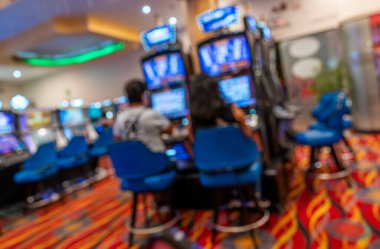 Renkli bir kumarhanede kumar oynayan insanların bulanık görüntüsü, parlak dijital ekranlar, mavi sandalyeler, ve yumuşak odaklı efektli canlı bir eğlence ortamı yaratan desenli halı..