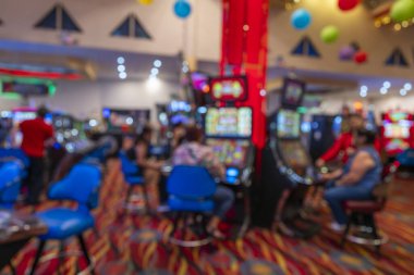 Parlak bir kumarhanede kumar oynayan insanların görüntüsü bulanık. Renkli dijital ekranlar, desenli halı ve mavi sandalyeler canlı, yumuşak odaklı bir eğlence ortamı yaratıyor..