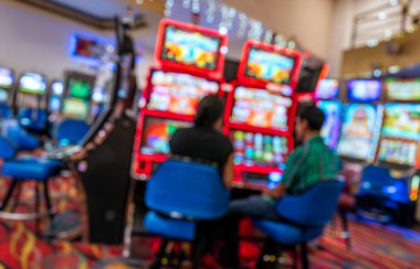 Kırmızı ve mavi ışıklandırma, dijital ekranlar, desenli halı ve yumuşak odak etkisiyle güçlendirilmiş canlı ortam ile parlak bir kumarhanede kumar oynayan iki kişinin bulanık sahnesi..