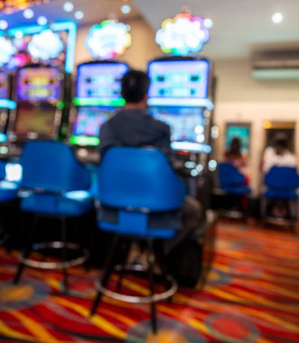 Parlak ışıklı bir kumarhanede kumar oynayan bir adamın bulanık sahnesi. Renkli ekranlar, desenli halı ve mavi sandalyeler canlı, yumuşak odaklı bir ortam yaratıyor..