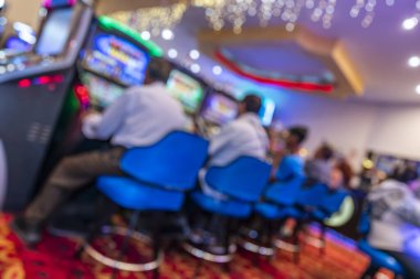 Mavi sandalyeli, kırmızı desenli halılı, neon ışıklı ve dekoratif tavan ışıklarıyla dolu renkli bir kumarhanede oturan oyuncuların bulanık bir görüntüsü. Ortam, kumar ve eğlence kültürünü yansıtıyor..