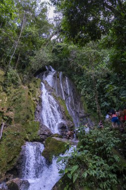 Peru yağmur ormanlarındaki büyük şelalede kayalık bölgeyi keşfeden ziyaretçiler var. Yoğun bitki örtüsü, akan su, yosunlu kayalar ve tropikal manzaralar seyahat ve macera temaları için ideal canlı bir doğal ortam yaratır..