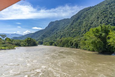 Ro Monzn, Peru Tingo Mara 'da yemyeşil dağların yanında akıyor. Parlak gökyüzü, yoğun bitki örtüsü ve canlı tropikal manzara seyahat, doğa ve açık hava temaları için ideal doğal bir manzara yaratır..