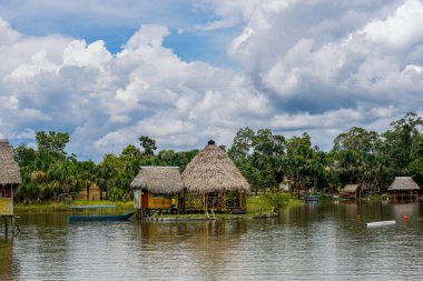 Geleneksel Amazon kulübeleri, Peru ormanlarında palmiye ağaçları ve yemyeşil bitki örtüsüyle çevrili sakin bir lagünün başında duruyor. Seyahat, doğa ve kültür temaları için barışçıl tropikal manzaralar yaratıyor..
