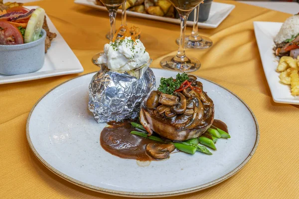 Mantar soslu fileminyon, kuşkonmaz ve folyoyla sarılmış kremalı patates, restoran ortamında seramik tabakta, gurme aromalı, zarif bir sunum, kaliteli bir yemek tarzı.