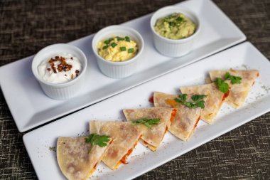Doldurulmuş quesadilla dilimleri, üç sosla birlikte dikdörtgen tabakta servis edilir. Gurme Meksika mezeleri restoran menüleri, yemek servisi, barlar ve günlük yemek kavramları için idealdir.