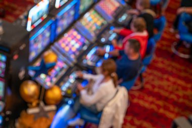 Kumarhane salonundaki kumar makinelerinde oynayan bulanık insanlar. Renkli ekranlar, parlak ışıklar ve dijital ekranlar kumar eğlence ve eğlence odaklı aktif bir atmosfer yaratıyor.