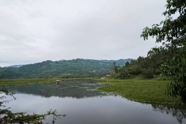 Yeşil tepeler ve yoğun bitki örtüsüyle çevrili huzurlu bir göl. Küçük tekneli sakin su, seyahat, turizm, ekoloji, sürdürülebilirlik, tatlı su ortamı ve açık hava manzarası için ideal kırsal manzarayı yansıtır.