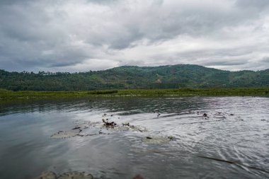 Yeşil tepeler ve yoğun bitki örtüsüyle çevrili huzurlu bir göl. Küçük tekneli sakin su, seyahat, turizm, ekoloji, sürdürülebilirlik, tatlı su ortamı ve açık hava manzarası için ideal kırsal manzarayı yansıtır.