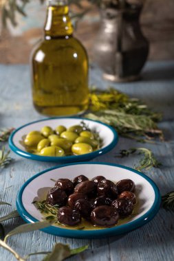 Mavi ahşap zemin üzerinde biberiye, fesleğen ve zeytin ağacı yapraklı doğal siyah ve yeşil zeytinler..