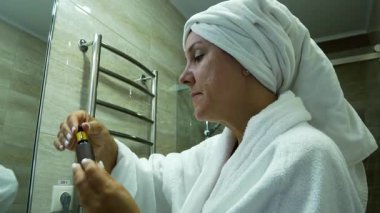 Banyoda, beyaz bornozlu ve kafasında havlu olan orta yaşlı bir kadın yüzüne kozmetik serumu sürüyor. Evde yüz bakımı..