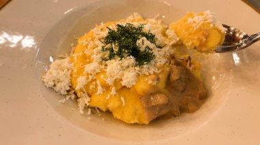 Mısır ezmesi banosh, keçi peyniri, otlar ve mantar sosu tabakta. Sahnenin arkasındaki kişi mısır gevreğini kaşıkla yer..