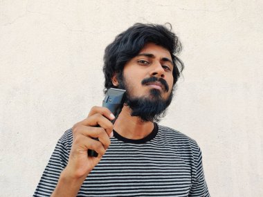 Sakallı, elinde makasla Güney Hintli genç bir adam. Uzun sakalını kesecek. Beyaz arka plan. Siyah tişört