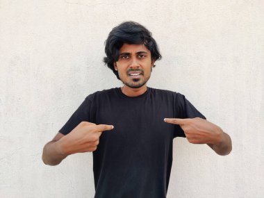 Gündelik görünüşlü genç Hintli Tamil adam kendini iki işaret parmağıyla işaret ediyor. Tişörtümü mü soruyorsun??