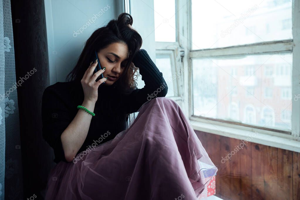 a depressed girl sits at the window with a phone in her hands on self-isolation
