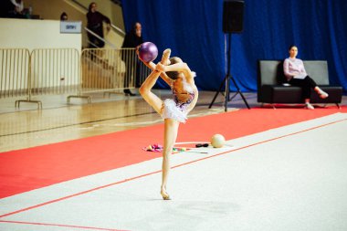 Bir kız artistik jimnastikçi bir yarışmada egzersiz yapıyor. Çevik sporcu güzel bir şekilde tek ayak üzerinde duruyor.