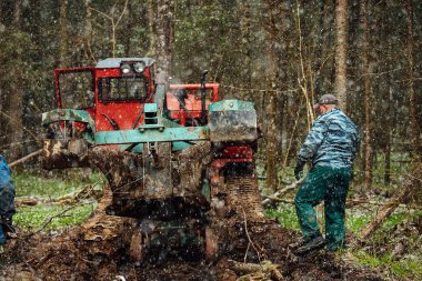 Bir sürüngen traktörü bir orman açıklığından geçiyor. Endüstriyel buldozer çamura saplanmış. Kamyonlar yerde kayıyor.