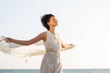 Beyaz elbiseli güzel bir kız elinde bir pareo ile deniz kıyısında yürüyor.