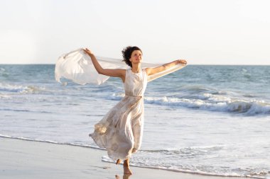 Beyaz elbiseli güzel bir kız elinde bir pareo ile deniz kıyısında yürüyor.
