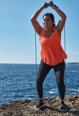 Latin kadın, orta yaşlı, spor giyer, spor giyer, antrenman yapar, fiziksel egzersiz yapar, kalas, mekik, tırmanış basamağı, kalori yakar, formda kalır, deniz kenarında, kulaklık takar, akıllı saat takar.