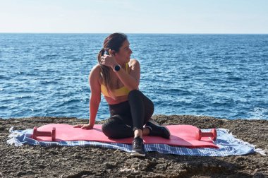 Latin kadın, orta yaşlı, spor giyer, spor giyer, antrenman yapar, fiziksel egzersiz yapar, kalas, mekik, tırmanış basamağı, kalori yakar, formda kalır, deniz kenarında, kulaklık takar, akıllı saat takar.