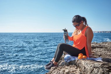Latin kadın, orta yaşlı, dinleniyor, gücünü geri kazanıyor, yemek yiyor, su içiyor, spor salonundan sonra, kalori yakıyor, formda, açık denizde, kulaklık takıyor ve akıllı saat takıyor.