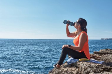 Latin kadın, orta yaşlı, dinleniyor, gücünü geri kazanıyor, yemek yiyor, su içiyor, spor salonundan sonra, kalori yakıyor, formda, açık denizde, kulaklık takıyor ve akıllı saat takıyor.