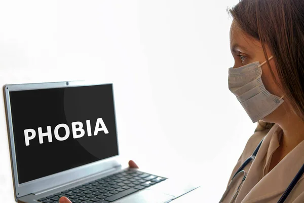 Medico donna in maschera con stetoscopio guarda lo schermo del monitor in cui è scritto il testo fobia — Foto Stock