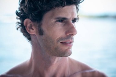Yakışıklı bir adam portresi, beyaz dişli üstsüz sörfçü gülüşü ve arka planda mavi okyanus.