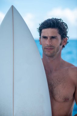Yakışıklı bir adamın portresi, beyaz sörf tahtası ve arka planda mavi okyanusu olan üstsüz bir sörfçü.
