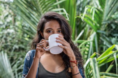 Genç esmer kadının portresi Bali 'de palmiye ağaçları manzaralı tropikal bungalovun balkonunda beyaz fincandan kahve içen serbest kadın.