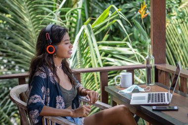Laptop, akıllı telefon ve kulaklıkla çalışan serbest çalışan genç esmer kadın portresi Bali 'deki palmiye ağaçları manzaralı tropik bungalovun balkonunda.