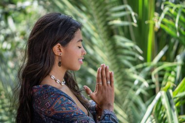 Genç esmer kadının portresi namaste pozisyonunda ellerini tropik bungalovun balkonunda, Bali 'de palmiye ağaçları manzaralı.