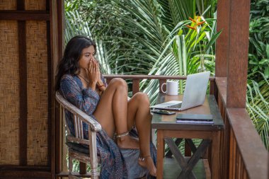 Genç esmer kadının portresi dizüstü bilgisayarla Bali 'deki tropik bungalovun balkonunda çalışıyor. Palmiye ağaçları manzaralı.