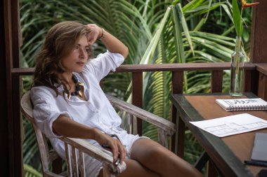 Genç, güzel, serbest çalışan bir kadının portresi. Bali 'de palmiye ağaçları manzaralı, tropik bungalovun balkonunda beyaz gömlekli bir defterle çalışıyor.