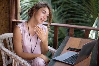Genç ve güzel bir kadının portresi dizüstü bilgisayarla Bali 'deki tropik bungalovun balkonunda çalışıyor. Palmiye ağaçları manzaralı.
