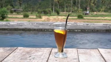 Bali 'deki pirinç tarlaları ve palmiye ağaçları için fevkalade bir manzarası olan küçük bir göletin kenarında bir bardak buzlu limon çayının video görüntüleri.