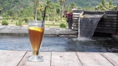 Bali 'deki pirinç tarlaları ve palmiye ağaçları için fıskiyeli küçük bir göletin kenarında bir bardak buzlu limon çayının video görüntüleri.