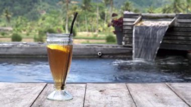 Bali 'deki pirinç tarlaları ve palmiye ağaçları için fıskiyeli küçük bir göletin kenarında bir bardak buzlu limon çayının video görüntüleri.