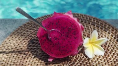 Taze kırmızı ejderha meyvesi, bir kaşıkla iki parçaya bölünmüş, köpüren mavi yüzme havuzunun kenarında, beyaz tropikal çiçekler, frangipani, Bali 'deki palmiye dalından gelen gölgeyi hareket ettiriyor.
