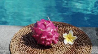 Video görüntüleri, taze kırmızı ejderha meyvesiyle yüzme havuzunun kenarında beyaz tropikal çiçeklerle frangipani 'yi, palmiye dalından gölgeyi ve Bali' de arka planda kaynayan mavi havuz suyunu gösteriyor. 